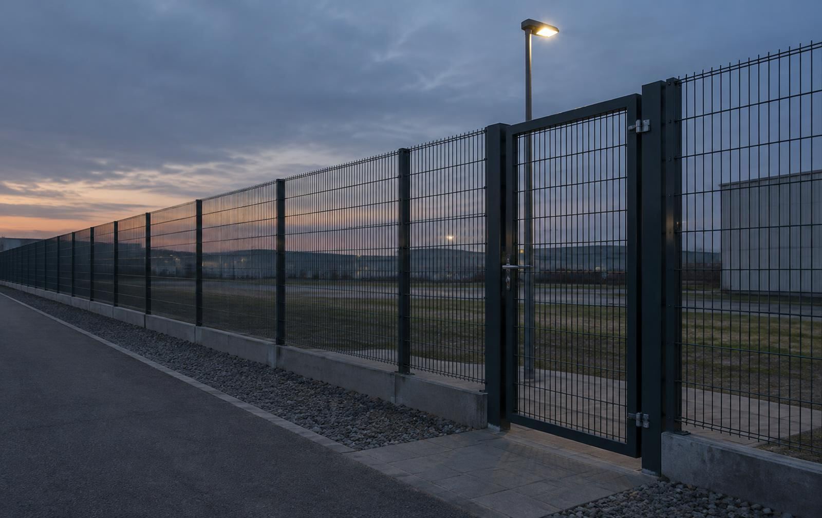 A long welded-mesh perimeter fence meeting a pedestrian access gate under a single pole light at dusk